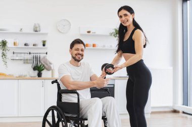 Engelli, olgun bir adamın karısıyla ya da kadın eğitmeniyle dambıl kullanırken çekilmiş portresi. Spor giyim sektöründe genç fizyoterapist ya da eş evde tekerlekli sandalye egzersizi yapan beyaz erkek hastaya yardım ediyor..