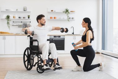 Kadın fizyoterapist ya da eş, tekerlekli sandalyedeki olgun erkek hastaya evde halterlerle nasıl egzersiz yapılacağını gösteriyor. Yaşlı hastalar için rehabilitasyon konsepti.