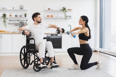 Kadın fizyoterapist ya da eş, tekerlekli sandalyedeki olgun erkek hastaya evde halterlerle nasıl egzersiz yapılacağını gösteriyor. Yaşlı hastalar için rehabilitasyon konsepti.