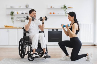 Yaşlı hastalar için rehabilitasyon konsepti. Kadın fizyoterapist ya da eş, tekerlekli sandalyedeki olgun erkek hastaya evde halterlerle nasıl egzersiz yapılacağını gösteriyor..