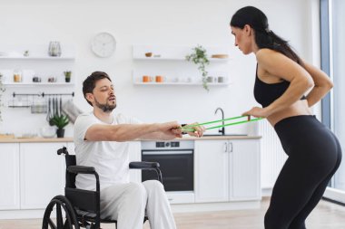 Spor giyim sektöründe genç bir fizyoterapist ya da eş evde tekerlekli sandalye egzersizi yapan erkek hastalara yardım ediyor. Engelli insanların rehabilitasyonu. Engelli, yetişkin, esneme bantlı adam eğitimi. .