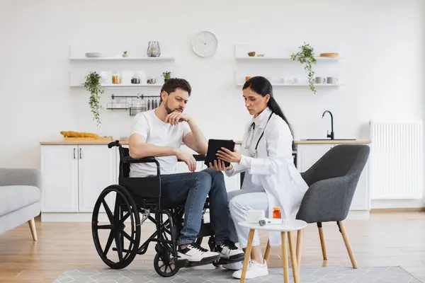 Full length shot of female brunette doctor checking up condition of bearded male patient at home. Nurse talking to mature man on wheelchair, showing examination results using modern tablet.
