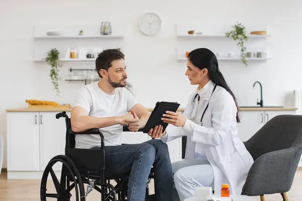 Portrait of brunette caucasian female doctor checking up condition of bearded male patient at home. Nurse talking to mature man on wheelchair, showing examination results using modern tablet.