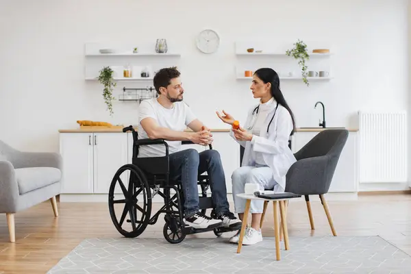 Kadın doktor, uzman kontrolleri yapıyor ve evde, mutfakta özürlü bir hastaya ilaç veriyor. Tıbbi geçmişle ilgili tavsiyeleri dinleyen odaklı tekerlekli sandalye kullanıcısı.