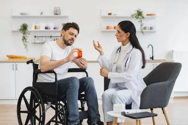 Tıbbi geçmişle ilgili tavsiyeleri dinleyen odaklanmış tekerlekli sandalye kullanıcısı. Kadın doktor, evde, mutfakta engelli bir hastaya uzman muayenesi ve ilaç veriyor..