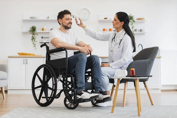 Kendine güvenen 30 yaşındaki kadın doktor ev ziyareti sırasında kızıl ötesi alın termometresi kullanarak tekerlekli sandalyedeki olgun sakallı hastanın vücut ısısını ölçüyor..