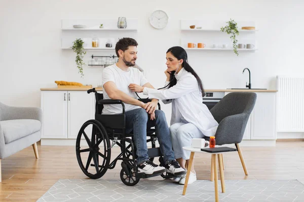 Beyaz önlüklü kadın steteskop kullanırken, yetişkin adam tekerlekli sandalyede oturuyor. Kalp atışlarını inceleyen pratisyen hekim evde düzenli kontrol sağlarken.