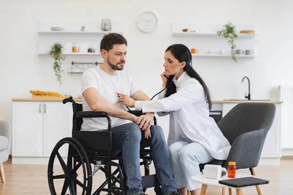 Beyaz önlüklü kadın steteskop kullanırken, yetişkin adam tekerlekli sandalyede oturuyor. Kalp atışlarını inceleyen pratisyen hekim evde düzenli kontrol sağlarken.