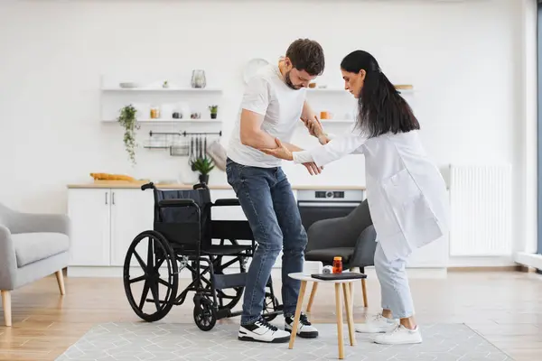 Fizikçi ona modern bir evde el uzatırken, tekerlekli sandalyenin yanında duran adamın tam boy görüntüsü. Başarılı bir tedaviden sonra hastayı engelli olarak teşvik eden tıbbi kadın..