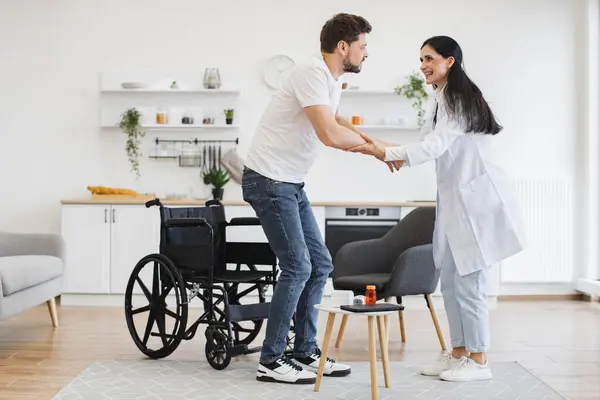 Başarılı bir tedaviden sonra hastayı engelli olmaya teşvik ediyor. Tekerlekli sandalyenin yanında duran adamın geniş açılı görüntüsü. Doktorlar ona modern evde el uzatıyor..