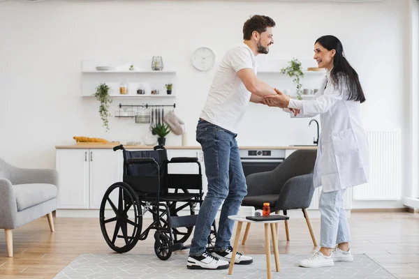 Beyaz tenli çekici bir kadının, engelli genç bir hastaya modern aydınlık mutfakta tekerlekli sandalyeden kalkması için yardım etmesi. Evdeki oturma odasında hasta destekleyen bir tıp uzmanı..
