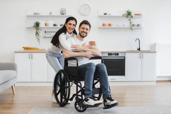 Portrait of young woman hugging Caucasian man with disability wearing casual outfit posing in spacious dining room. Positive young husband feeling optimistic while making recovery from injury at home.