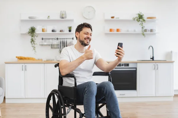 Smiling Caucasian man chatting online with relatives via video call application in dining room. Joyful young adult with disability holding mobile while planning outdoor travel in modern apartment.