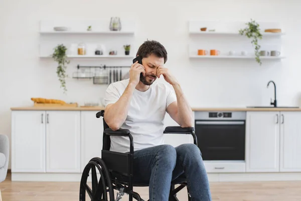 Tired person discussing after dinner activity with friends outside apartment. Close up view of bearded male with mobility impairment being involved in phone talk while relaxing in home interior.