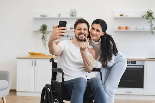 Wheelchair user and his wife in casual outfit using phone webcam while enjoying free time. Young family with disability receiving online video call via mobile application and speaking with relatives.