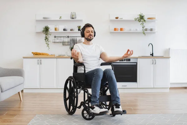 Portrait of smiling caucasian gentleman in headphones resting in wheelchair in lotus position on kitchen background. Relaxed mature adult with disability promoting healthy lifestyle via meditation.