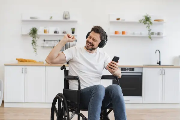 Cheerful Caucasian adult dancing along while hearing loud tunes in headphones. Joyous man in white t-shirt raising hand while looking at smartphone sitting in wheelchair in studio apartment.