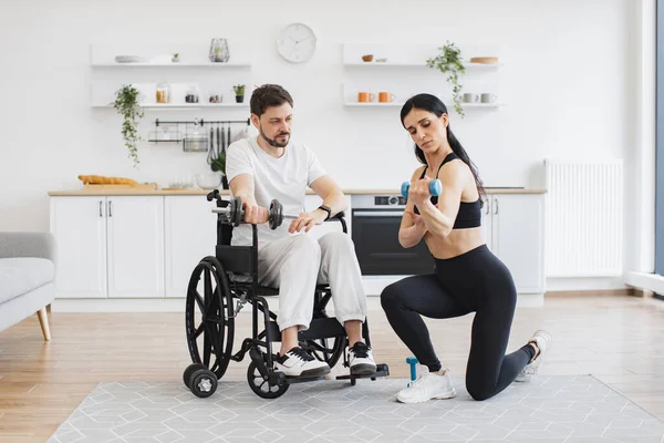 Genç erkek hasta için rehabilitasyon. Spor giyim sektöründe milenyum fizyoterapisti tekerlekli sandalyedeki engelli bir adamın halterlerle antrenman yapmasına yardım ediyor..