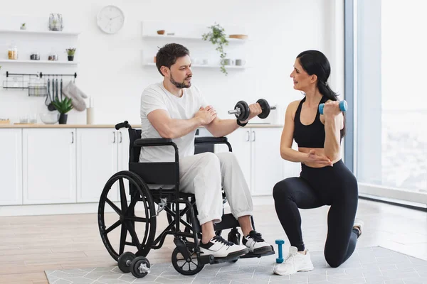 Erkek hasta, modern bir apartmanın arka planında hastalık ve yaralanma üzerine rehabilitasyona giriyor. Spor salonundaki fizyoterapist, tekerlekli sandalyedeki yetişkin bir adama evde dambıllarla nasıl egzersiz yapıldığını gösteriyor..