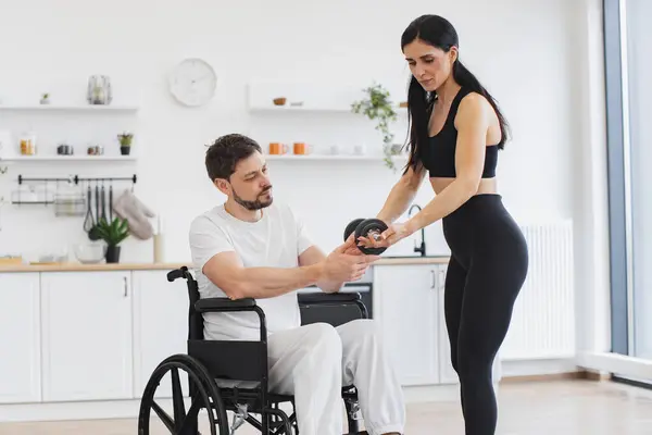 Rehabilitation of disabled people concept. Young physiotherapist or wife in sportswear helping Caucasian male patient in wheelchair exercise at home. Handicapped mature man training with dumbbells.