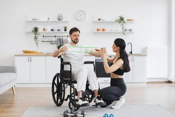Concept of rehabilitation of disabled people. Young physiotherapist or wife in sportswear helping male patient in wheelchair exercise at home. Handicapped mature man training with stretching bands .
