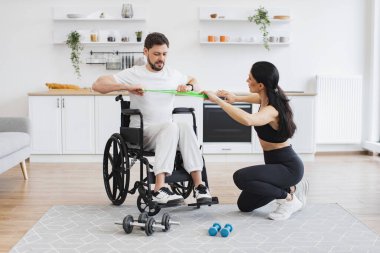 Engelli, yetişkin, esneme bantlarıyla antrenman yapıyor. Engelli insanların rehabilitasyonu. Spor giyim sektöründe genç fizyoterapist ya da eş evde tekerlekli sandalye egzersizi yapan erkek hastalara yardım ediyor..