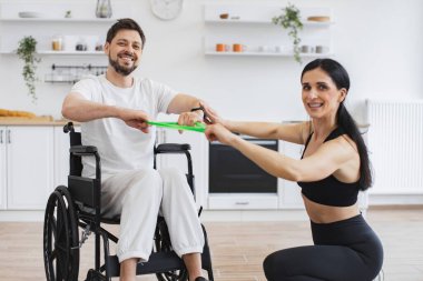Rehabilitasyon sırasında kadın fitness eğitmeni ve erkek hasta portresi. Fizyoterapist tekerlekli sandalyedeki engelli bir adama esneme bantlarıyla çalışması için yardım ediyor, evde iyileşmesi için.
