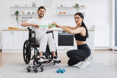 Rehabilitasyon sırasında kadın fitness eğitmeni ve erkek hasta portresi. Fizyoterapist tekerlekli sandalyedeki engelli bir adama esneme bantlarıyla çalışması için yardım ediyor, evde iyileşmesi için.