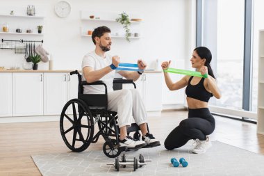 Kadın fizyoterapist ya da eş tekerlekli sandalyedeki olgun erkek hastaya evde esneme bantlarıyla nasıl egzersiz yapılacağını gösteriyor. Yaşlı hastalar için rehabilitasyon konsepti.