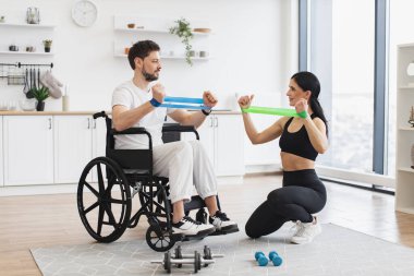 Kadın fizyoterapist ya da eş tekerlekli sandalyedeki olgun erkek hastaya evde esneme bantlarıyla nasıl egzersiz yapılacağını gösteriyor. Yaşlı hastalar için rehabilitasyon konsepti.