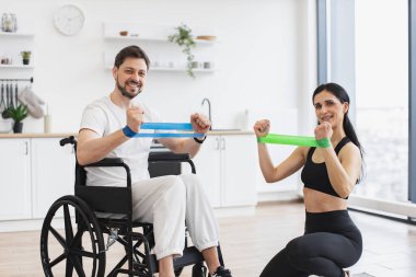 Kadın fizyoterapist portresi ve tekerlekli sandalyedeki adam evde esneme bantlarıyla egzersiz yapıyorlar. Erkek hasta, modern apartmanın arka planında hastalık ve yaralanma üzerine rehabilitasyon görüyor