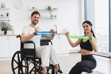 Kadın fizyoterapist portresi ve tekerlekli sandalyedeki adam evde esneme bantlarıyla egzersiz yapıyorlar. Erkek hasta, modern apartmanın arka planında hastalık ve yaralanma üzerine rehabilitasyon görüyor