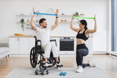 Spor kıyafetli bir eş, tekerlekli sandalyedeki olgun bir adama kas gücü için kollarını gerdirmesine yardım ediyor. Fizyoterapi sağlık hizmetleri, tıbbi eğitmen engelli beyaz hastalara danışmanlık yapıyor..