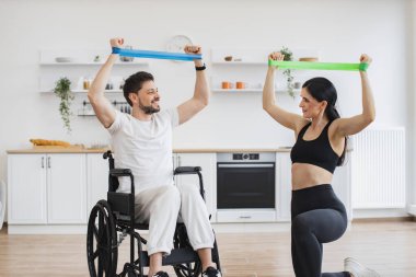 Fizyoterapi sağlık hizmetleri, tıbbi eğitmen engelli beyaz hastalara danışmanlık yapıyor. Spor kıyafetli eş, tekerlekli sandalyedeki olgun bir adama kas gücü için kollarını gerdirerek yardım ediyor..