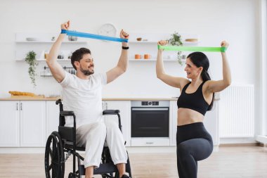 Fizyoterapi sağlık hizmetleri, tıbbi eğitmen engelli beyaz hastalara danışmanlık yapıyor. Spor kıyafetli eş, tekerlekli sandalyedeki olgun bir adama kas gücü için kollarını gerdirerek yardım ediyor..