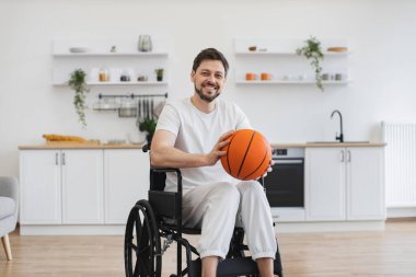 Tekerlekli sandalyedeki çekici beyaz erkek portresi. Apartman mutfağında basketbolla poz veriyor. Gülümseyen sakallı insan içeride fiziksel aktivitenin gerçek sağlığına kavuşuyor..
