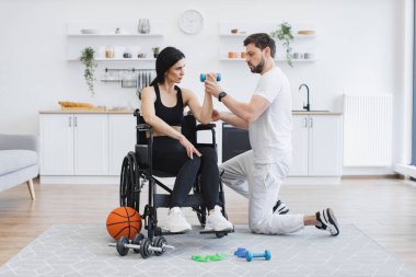 Spor giyim sektöründe fizyoterapist ya da koca evde tekerlekli sandalye egzersizi yapan beyaz kadın hastalara yardım ediyor. Engelli insanların rehabilitasyonu. Engelli, olgun kadın eğitiminde..