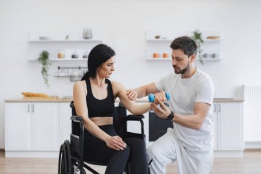 Engelli, olgun, halterli kadın eğitimi. Spor giyim sektöründe fizyoterapist ya da koca evde tekerlekli sandalye egzersizi yapan beyaz kadın hastalara yardım ediyor. Engelli insanların rehabilitasyonu.