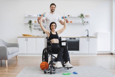 Erkek fizyoterapist ya da koca tekerlekli sandalyedeki olgun kadın hastaya evde halterlerle nasıl egzersiz yapacağını gösteriyor. Yaşlı hastalar için rehabilitasyon konsepti.