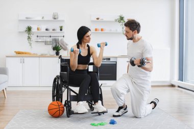 Spor giyim sektöründe milenyum fizyoterapisti tekerlekli sandalyedeki engelli yetişkin bir kadına halterlerle çalışması için yardım ediyor, evdeki sakatlıktan kurtulması için. Genç bayan hastalar için rehabilitasyon.