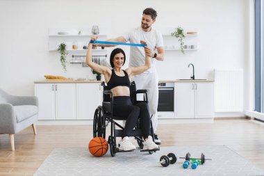 Spor kıyafetli koca, tekerlekli sandalyedeki olgun bir kadının kas gücü için kollarını germesine yardım ediyor. Fizyoterapi sağlık hizmetleri, tıbbi eğitmen engelli beyaz hastalara danışmanlık yapıyor..