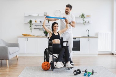 Spor kıyafetli koca, tekerlekli sandalyedeki olgun bir kadının kas gücü için kollarını germesine yardım ediyor. Fizyoterapi sağlık hizmetleri, tıbbi eğitmen engelli beyaz hastalara danışmanlık yapıyor..