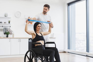 Fizyoterapi sağlık hizmetleri, tıbbi eğitmen engelli beyaz hastalara danışmanlık yapıyor. Spor giyimli koca, tekerlekli sandalyedeki olgun bir kadına kas gücü için kollarını gerdirerek yardım ediyor..