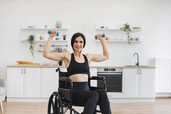 Joyful Caucasian person with physical disability holding dumbbells in raised arms during strength training at home. Active wheelchair user building muscles while experimenting with workout routine.