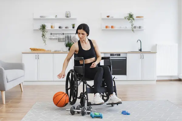 Cheerful beautiful woman with mobility impairment dribbling ball in studio apartment on weekend. Happy Caucasian in black sportswear staying active with disability while playing game for pleasure.