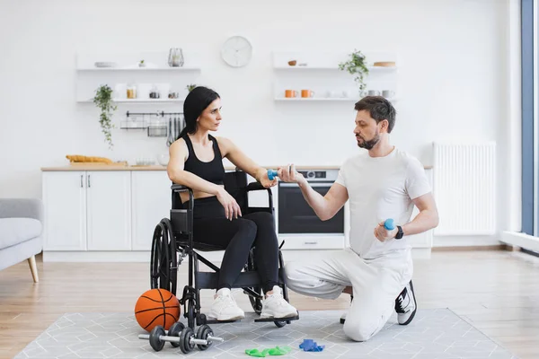 Rehabilitation of disabled people concept. Physiotherapist or husband in sportswear helping Caucasian female patient in wheelchair exercise at home. Handicapped mature woman training with dumbbells.