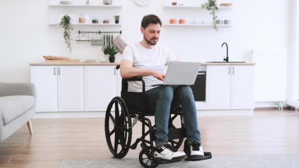 明るいオープンプランのキッチンに滞在しながら 膝の上にラップトップを持っている車椅子の楽しい白人男性 幸せな若い大人のサーフィンインターネットウェブページ自宅での事故後に回復しながら — ストック動画