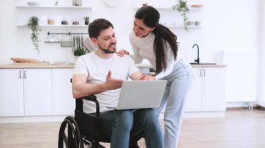 Evde geçirdiğin kazadan sonra internet sitelerinde mutlu sörfler. Tekerlekli sandalyedeki beyaz adam dizlerinin üzerinde dizlerinin üzerinde karısı için parlak mutfağın ortasında iş sonuçlarını gösteriyor..