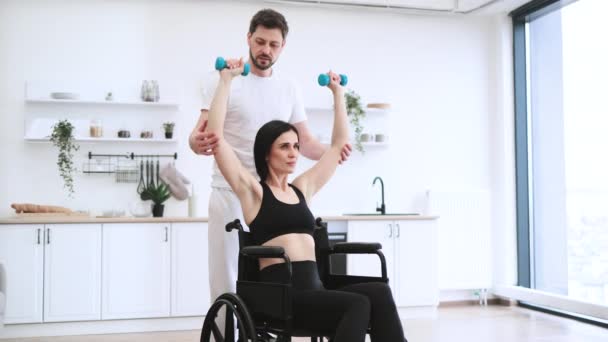 医学療法のヘルスケア 医師のコンサルティングは 慎重な患者を無効にしました リハビリテーションの筋力のためのダンベルで腕を持ち上げる車椅子の成熟した女性を助けるトレーナーまたは夫 — ストック動画