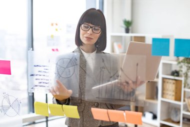 Modern teknoloji kavramı ve meşgul insanlar. Resmi giyinmiş, gözlüklü, kendine güveni tam beyaz bir kadın şirket yönetim kurulunda dikilirken dizüstü bilgisayarla iş sorunları çözüyordu..